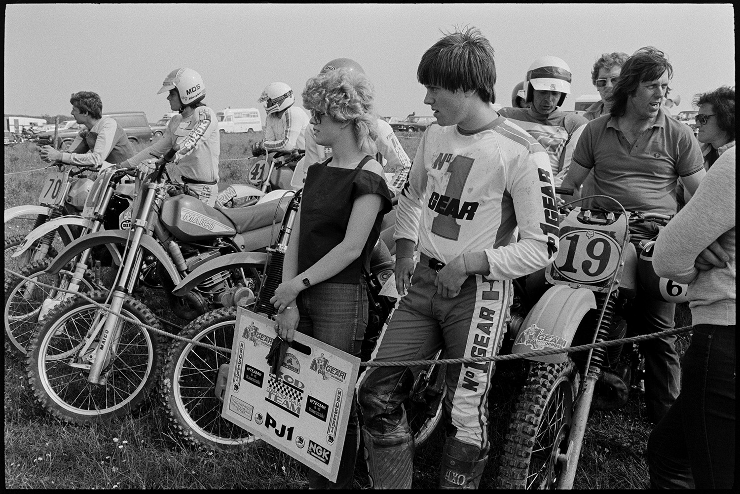 Motorcycle scramble competitors waiting to start racing and jumping at start line. 
[Competitors waiting at the start line of a motorcycle scramble race in a field at Torrington. A woman is stood at the front holding a placard.]