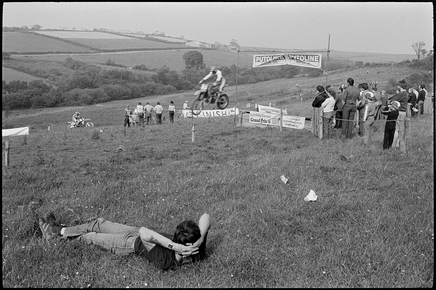 Motorcycle scramble competitors waiting to start racing and jumping at start line. 
[Spectators watching competitors jumping a ramp at the start of a motorcycle scramble in a field at Torrington. Other competitors can be seen racing around the course in the background.]