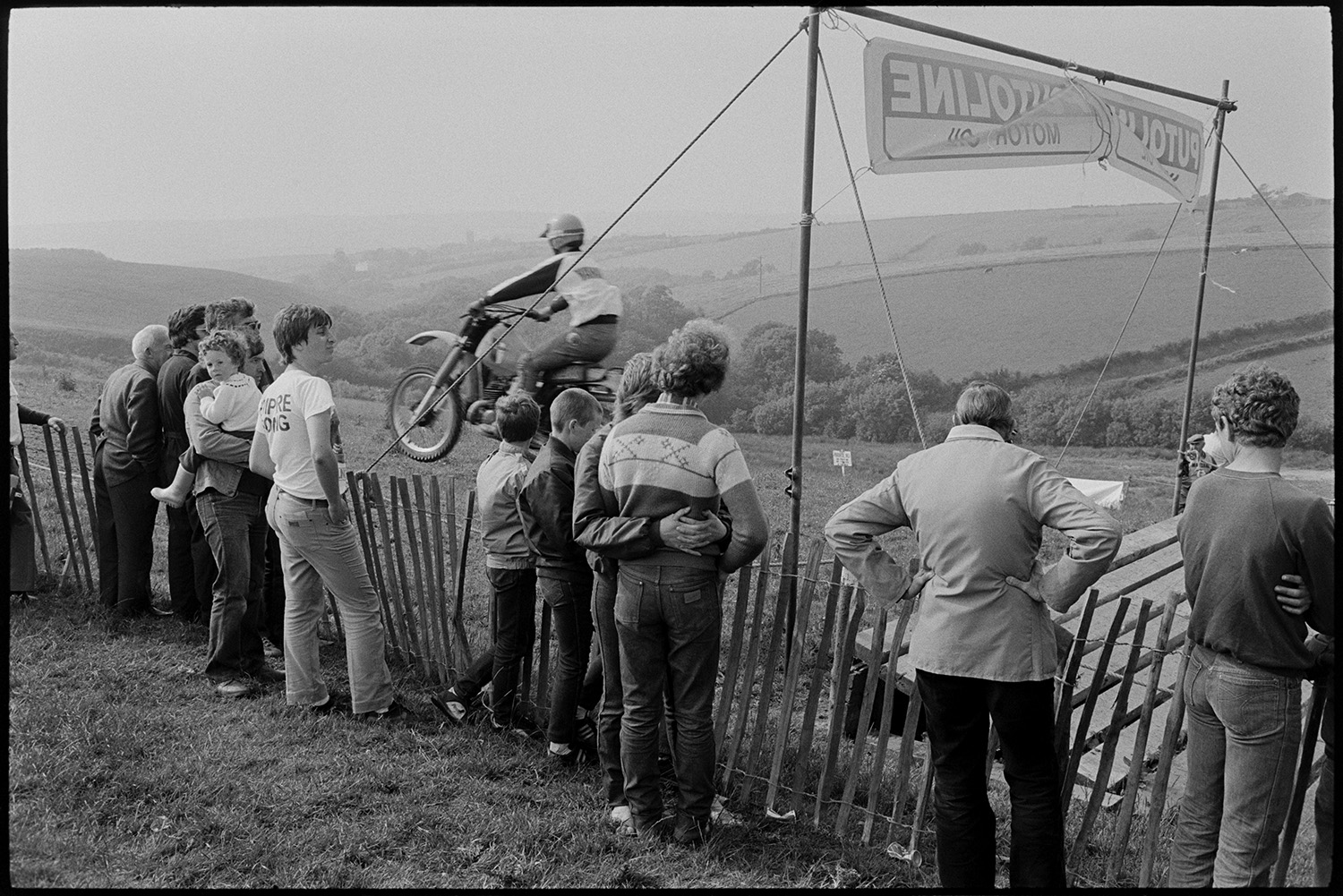 Motorcycle scramble competitors waiting to start racing and jumping at start line. 
[Spectators, including children, watching competitors jumping off a ramp at the start of a motorcycle scramble race in a field at Torrington.]