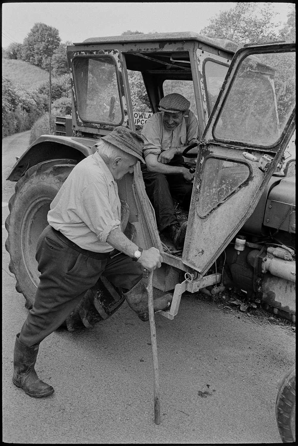 Farmers chatting in road, dog and tractor. 
[Archie Parkhouse leaning on a tractor and walking stick, talking to George Ayre who is sat in the cab of the tractor, in a lane at Millhams, Dolton.]