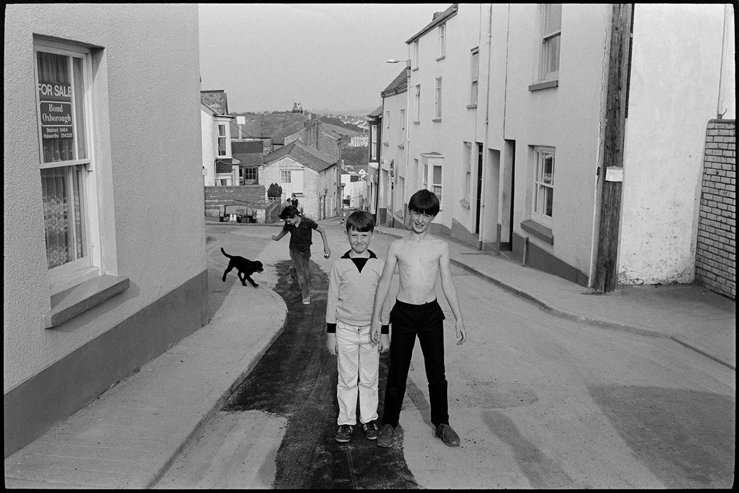 Street scenes, shops, passers by, woman cleaning front of house sweeping. 
[Three boys and dog playing in a street on a hill in Bideford. The house next to two of the boys is for sale. Part of the road has recently been tarmacked.]