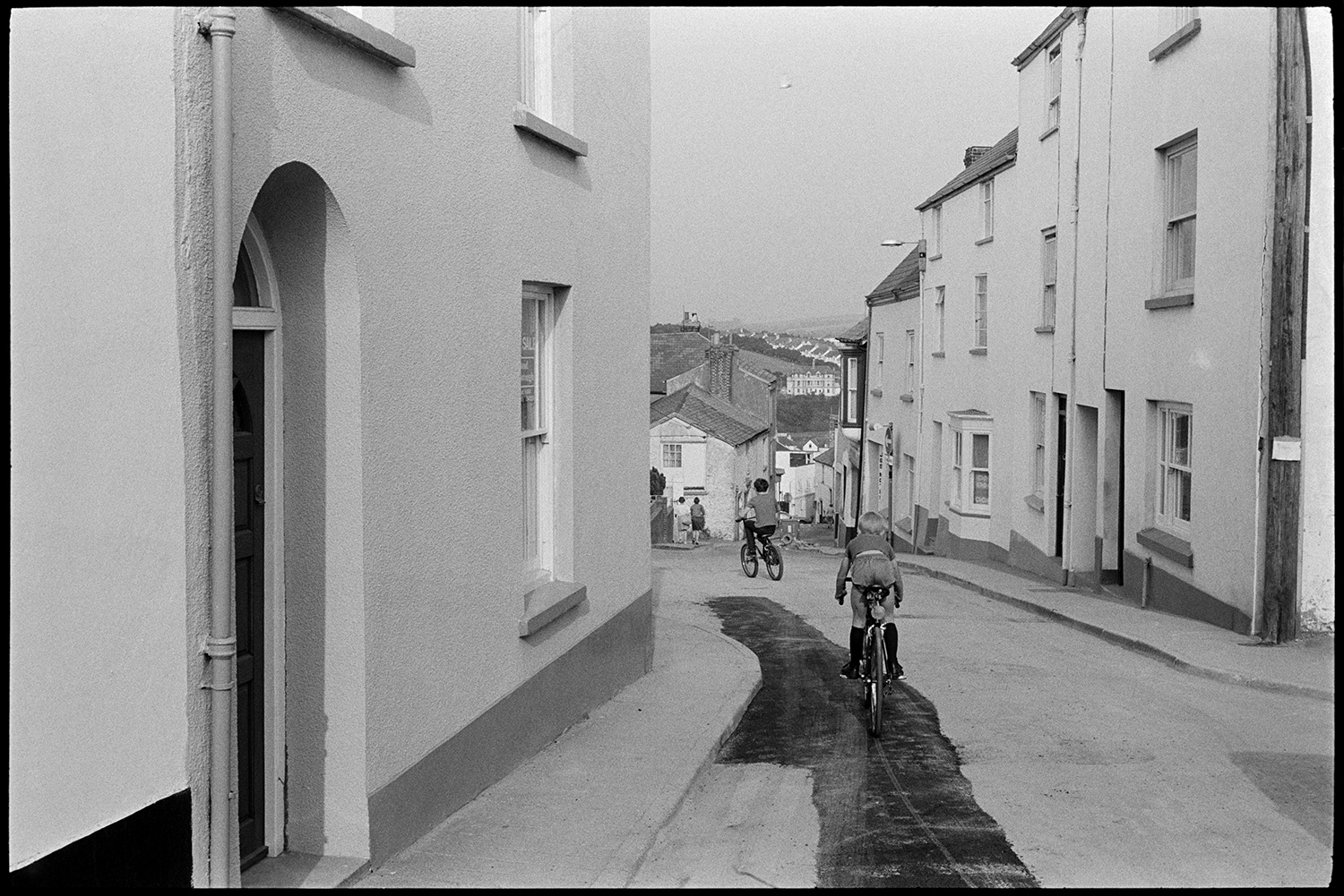 Street scenes, shops, passers by, woman cleaning front of house sweeping. 
[Two children riding bicycles past houses down a street on a hill in Bideford. Part of the road has recently been tarmacked.]