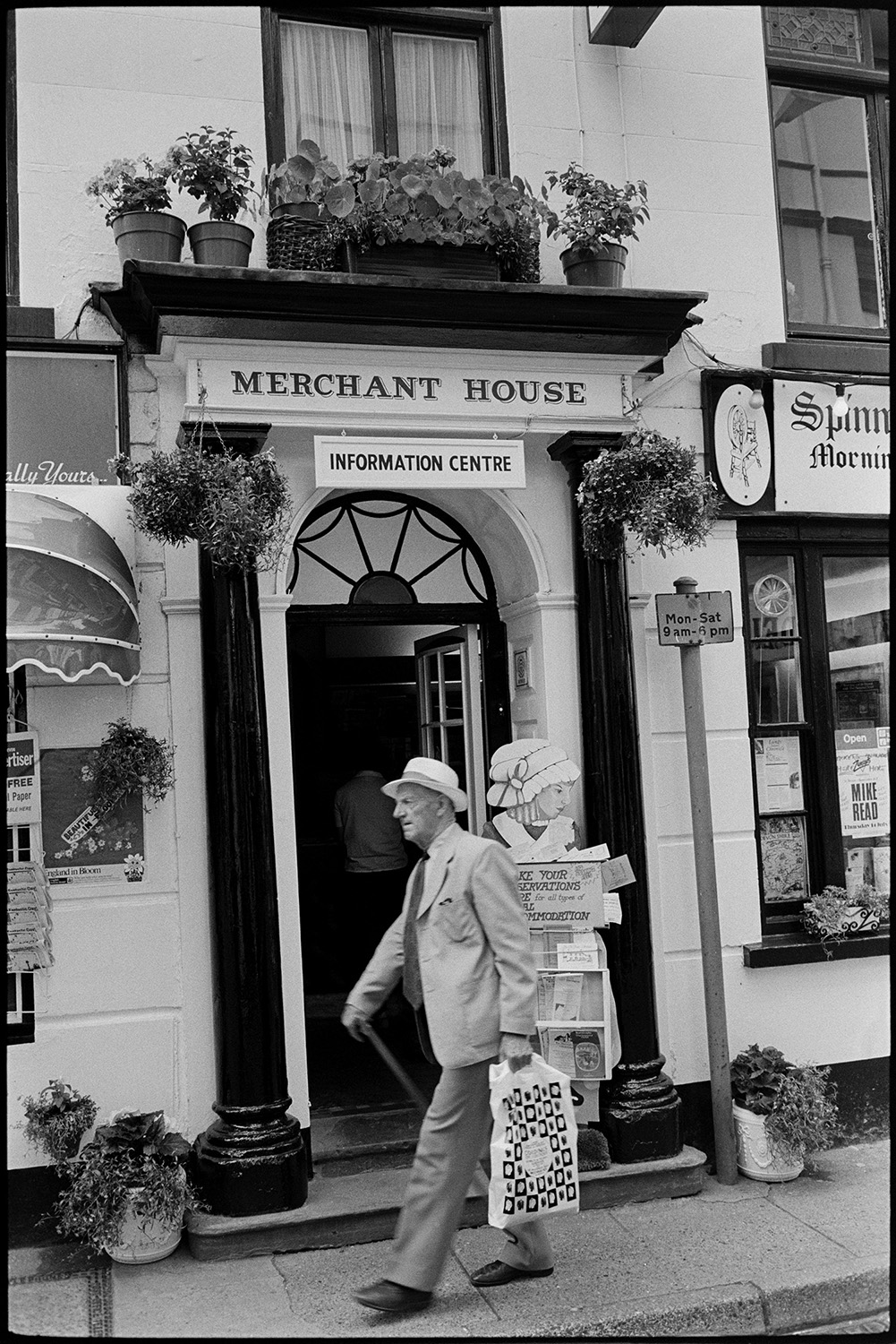 Doorways and shopfronts. 
[A man wearing a straw hat, carrying a bag and holding a walking stick, passing the entrance to the tourist information centre at Merchant House in Bideford. The doorway is decorated with plant pots and hanging baskets.]