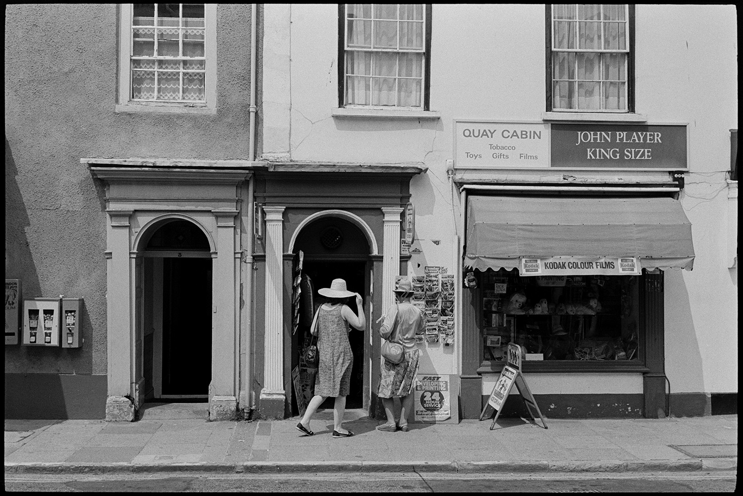 Doorways and shopfronts. 
[Two women, wearing straw hats, looking at postcards outside the Quay Cabin shop front in Bideford.]