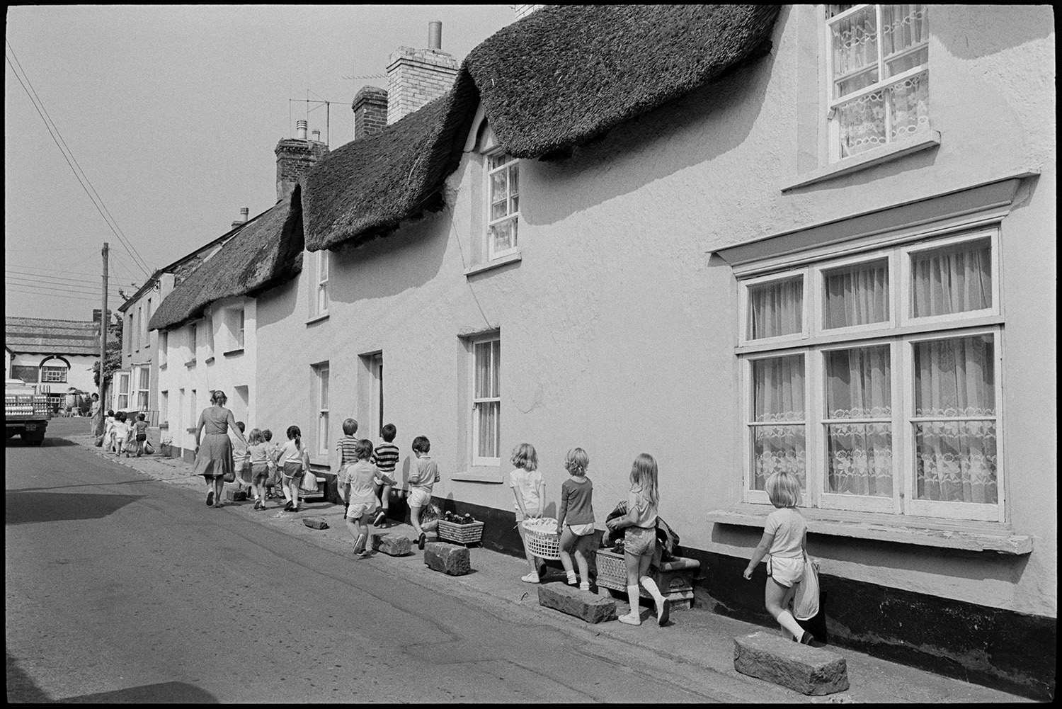 Street scene, schoolchildren going back to school after sports. 
[A teacher and group of school children walking past thatched cottages in Fore Street, Dolton, on their way back to school after a sports lesson. Some of the children are carrying baskets with sports equipment.]