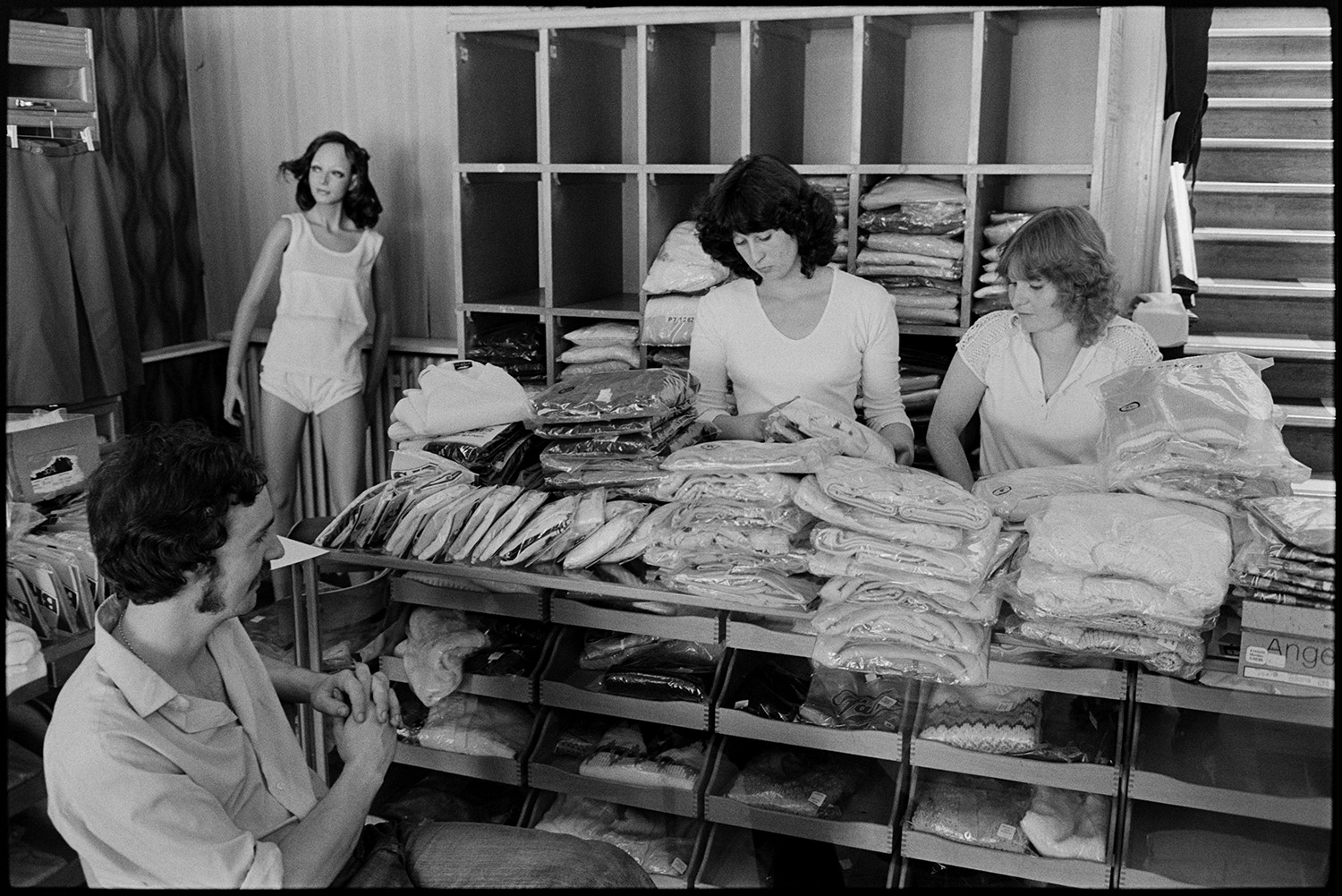 Women serving behind counter of clothes shop, shelves and dummy, chatting with customer. 
[Two women working in a clothes shop in Buttgarden Street, Bideford. They are sorting packets of clothing while talking to a man sat in front of the counter. A shop dummy or mannequin and shelves with more clothes can be seen in the background.]