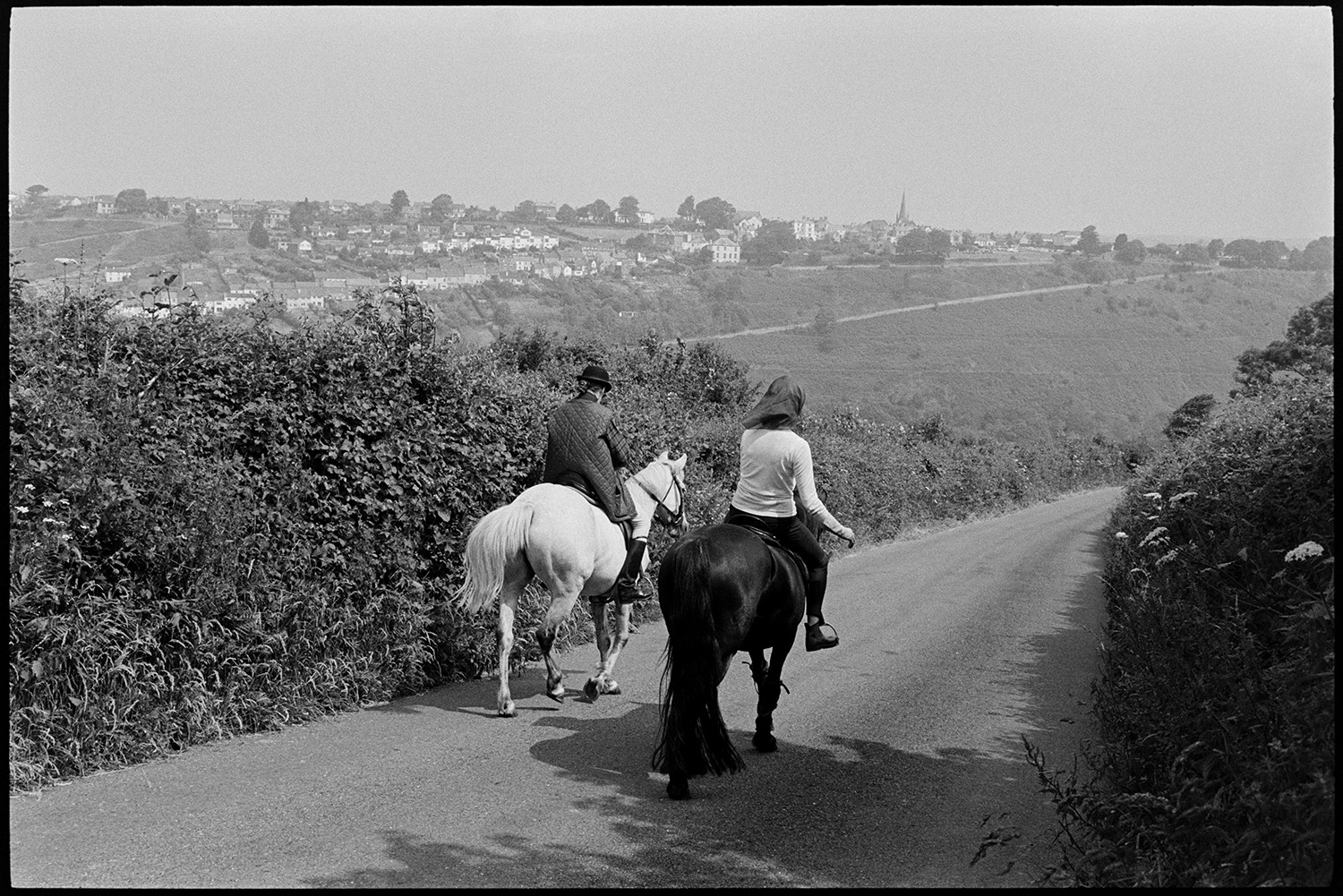 Two horses in road. 
[Two people riding horses along a hedge lined road near Torrington. The town of Torrington can be seen in the distance.]