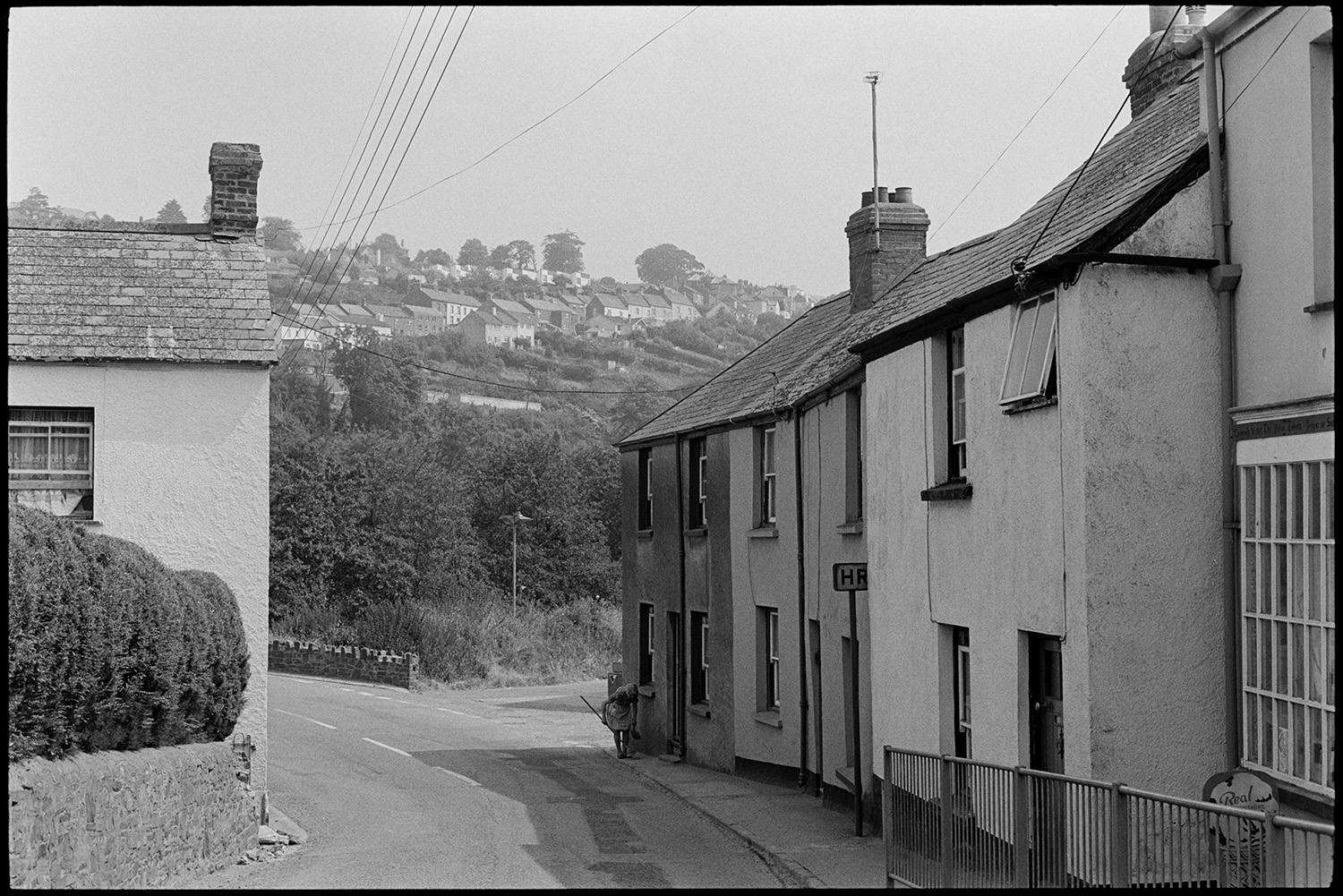 Row of cottages. 
[A road with terraced cottages in Little Torrington. A woman is sweeping outside one of the cottages. Another row of cottages can be seen on a hill in the background.]