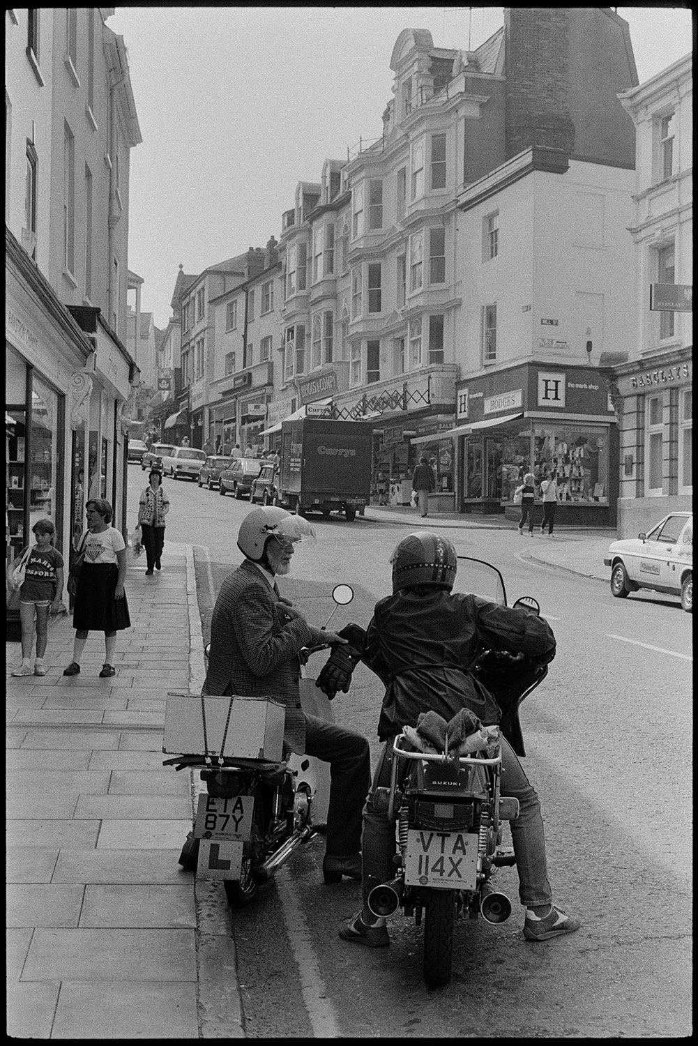 Street scenes, motorbikes, car. 
[Two people talking whilst sat on motorbikes parked on the side of the High Street in Bideford. Shoppers, shop front and other parked cars can be seen further along the street.]