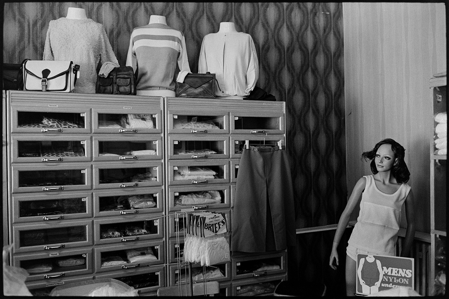 Interior of clothes shop shelves and displays of goods. Woman doing accounts, staircase. 
[Clothes displayed in shelves or display cases in a clothes shop in Buttgarden Street, Bideford. On top of the shelves is a display of bags and jumpers. A mannequin or shop dummy is stood next to the shelves.]