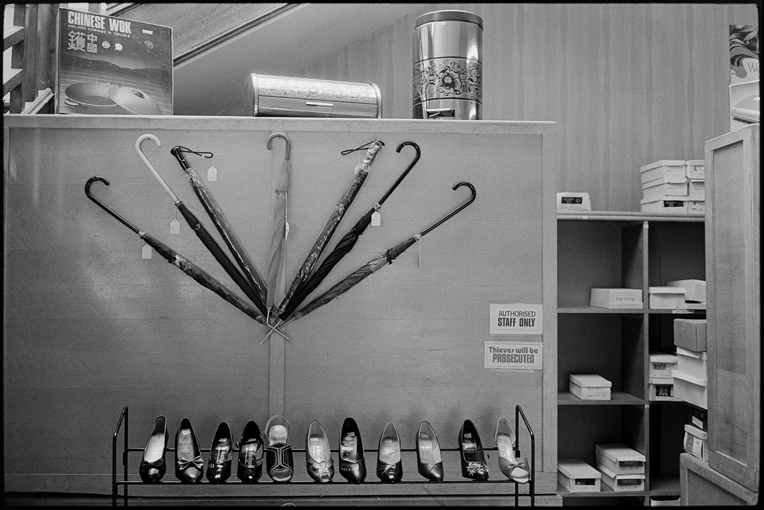 Interior of clothes shop shelves and displays of goods. Woman doing accounts, staircase. 
[A display of shoes and umbrellas in a clothes shop in Buttgarden Street, Bideford. A Chinese wok, bread bin and flip bin are displayed on top of the cabinet with the umbrellas. Shoe boxes can also be seen on shelves in the background.]