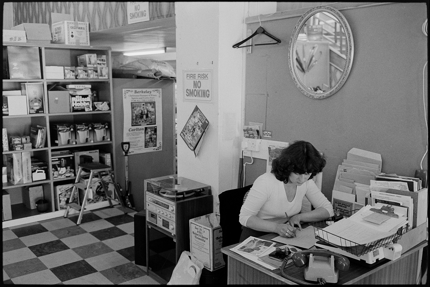 Interior of clothes shop shelves and displays of goods. Woman doing accounts, staircase. 
[A woman working on accounts at a desk in a shop in Buttgarden Street, Bideford. Various files and a telephone are on the desk in front of her. Goods are displayed on shelves in the background, including kettles and a food processor. A record player is also visible behind her desk.]