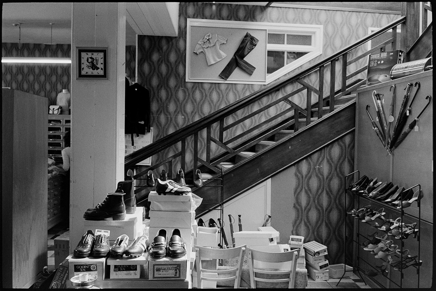 Interior of clothes shop shelves and displays of goods. Woman doing accounts, staircase. 
[A display of shoes and umbrellas below a staircase in a shop in Buttgarden Street, Bideford. The wall behind the staircase is covered with patterned wallpaper and a display of a pair of trousers and a top.]