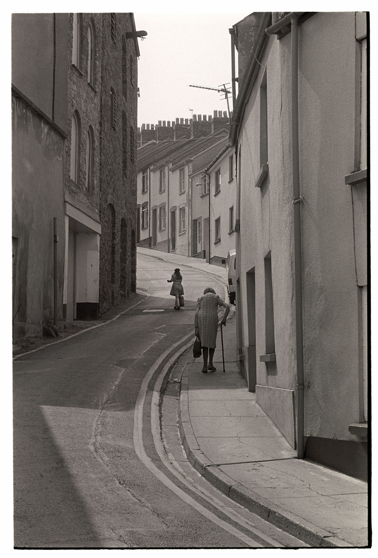 Street scene, elderly woman with stick climbing hill. 
[Two women walking up a street on a hill with a row of terraced houses at Bideford. One of the women is using a walking stick and carrying a bag.]