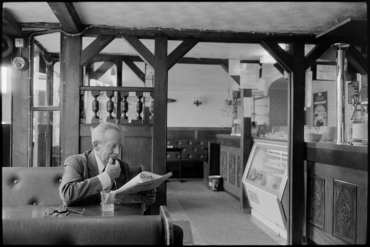 Man having drink and reading paper in cafe, restaurant. 
[A man reading a newspaper and having a drink at a table in a café in Mill Street, Bideford. The interior of the café is visible with a wooden panelled counter and wooden beams.]