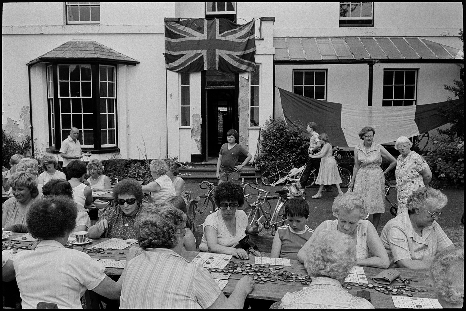 Vicarage fete, stalls, women playing bingo on lawn, teas and cakes. 
[Women and children playing bingo at a fete at Torrington Vicarage. Women and a child are watching in the background by bicycles. A couple pushing a pram and a man holding a cup of tea are also visible. The vicarage buildings is decorated with flags, including the Union Jack flag.]