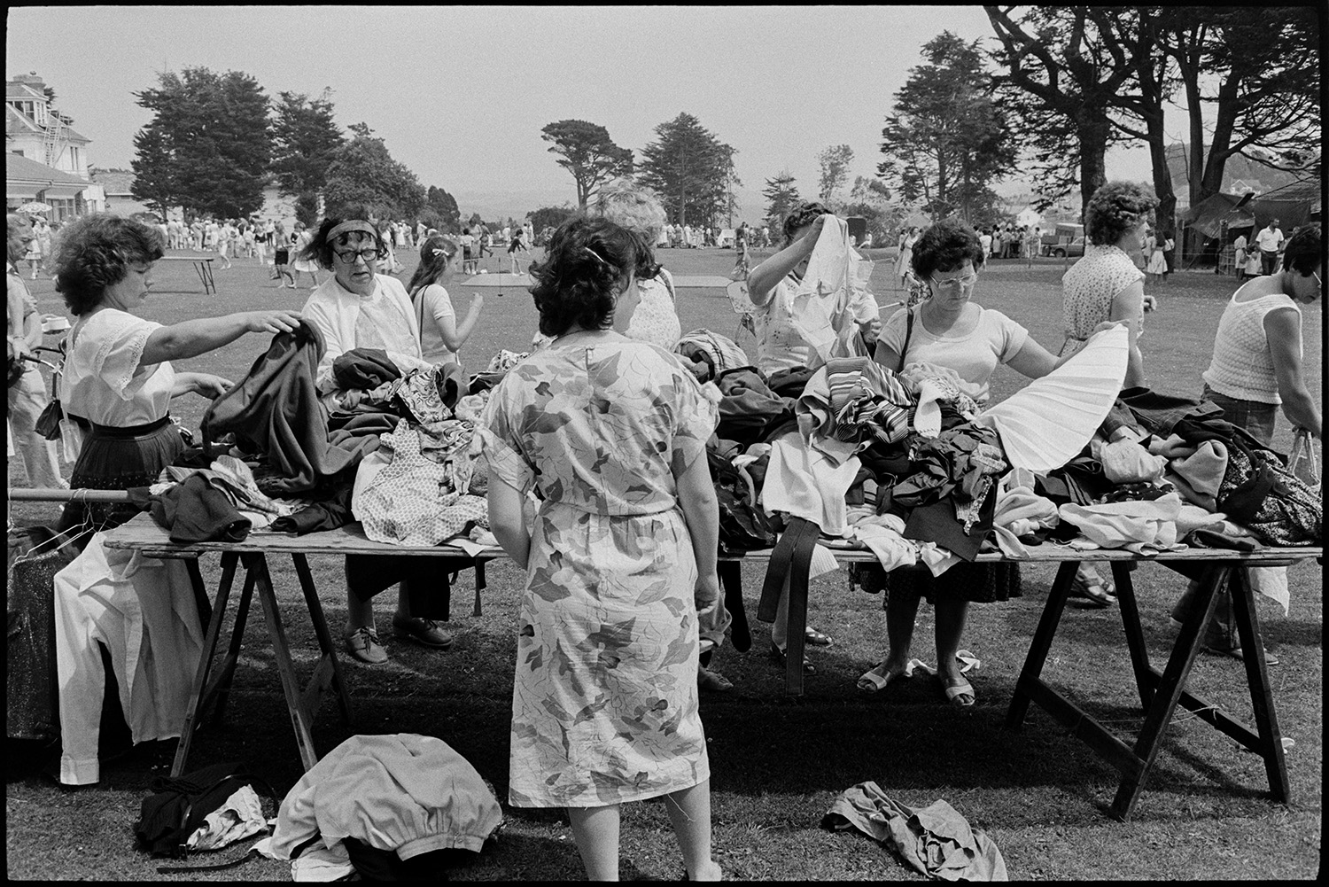 School fete. Stalls. Fancy dress. Pets being judged, pet slug 22! Produce, dogs. Vintage Car rides. 
[Women looking through clothes on a jumble stall at the school fete at Edgehill College, Bideford. Other stalls and people can be seen in the background on the school field.]