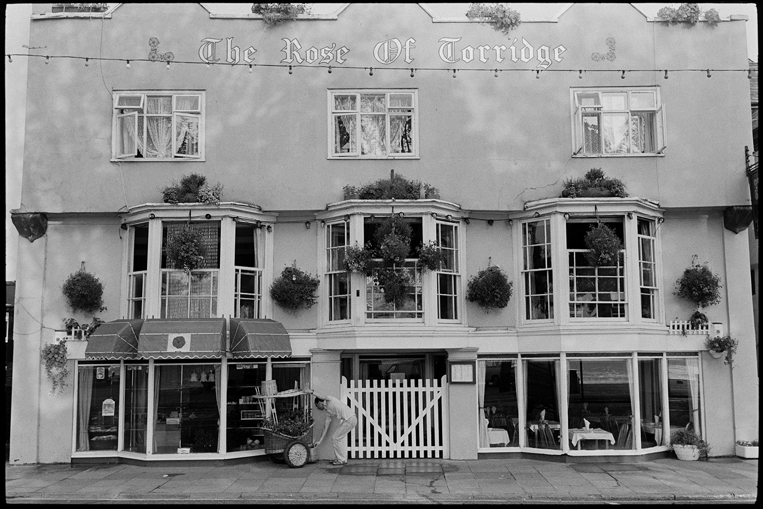 Front of Restaurant, Tea Room. 
[The shop front of The Rose of Torridge tea rooms or restaurant at Bideford Quay. A person is securing a small wagon outside with plants and the windows are decorated with hanging baskets.]