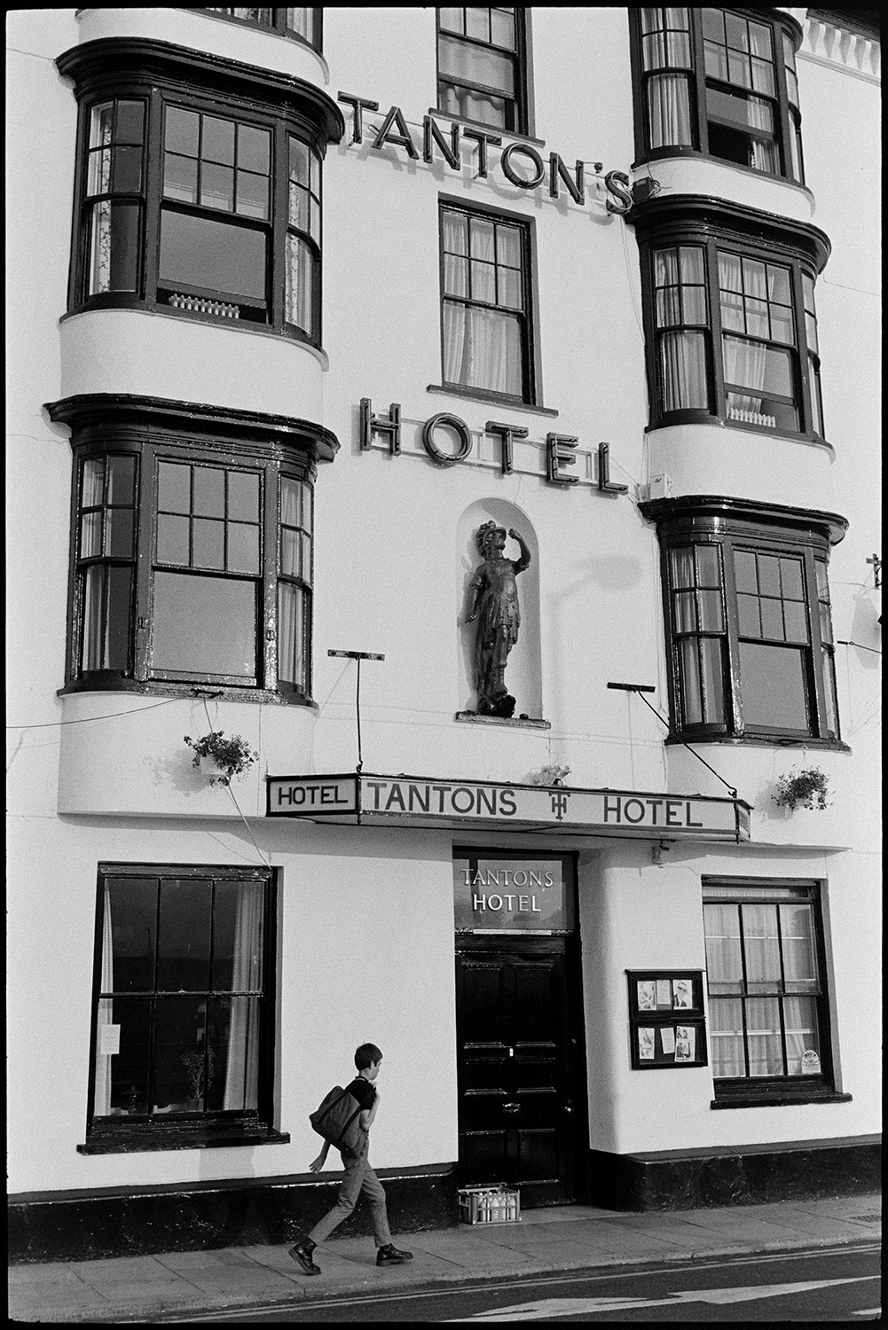 Front of hotel, with signs. 
[A person walking past the front of Tanton's Hotel at Bideford. A statue is above the hotel sign.]