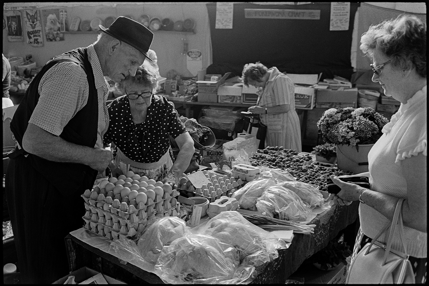 Pannier market stalls, cheese eggs, vegetables, customers. 
[A woman buying eggs from a man and woman running a stall at Bideford Pannier Market. Poultry, punnets of fruit and flowers are also for sale on the stall.]