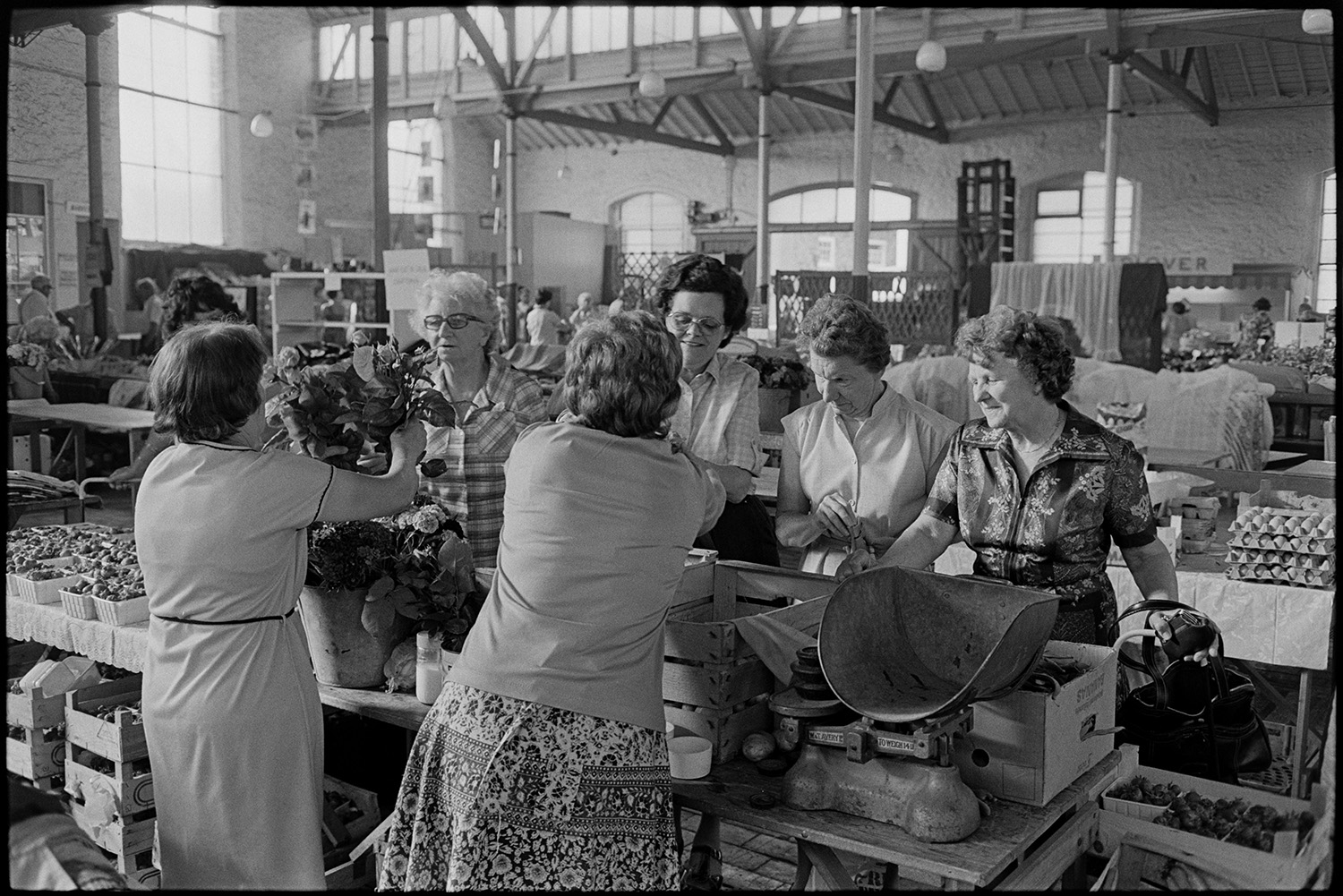 Pannier market stalls, cheese eggs, vegetables, customers. 
[Women buying flowers and vegetables from two women running a stall at Bideford Pannier Market. A set of scales can be seen on the stall. Other stalls are visible in the background, including one with eggs.]