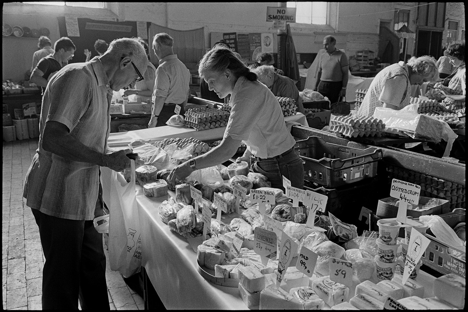 Pannier market stalls, cheese eggs, vegetables, couple setting up stall with eggs in baskets. 
[A man buying cuts of meat from a woman running a stall at Bideford Pannier Market. The stall is also selling cheese and clotted cream. Other stalls, including one selling eggs can be seen in the background.]