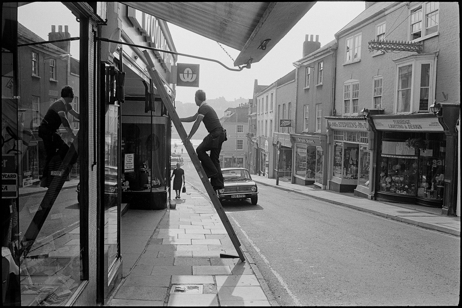 Window cleaner up ladder in front of shop in high street, early morning sun. 
[A man climbing a ladder to clean a shop window in Bideford High Street. Other shop fronts can be seen along the street, including Torridge Lighting and Design, Stephens, Briggs Shoes and Boots.]