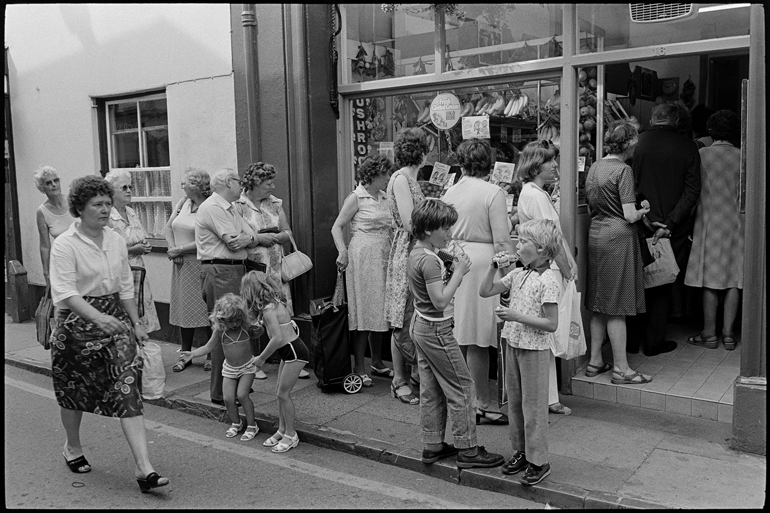 Street scenes, passers by. 
[Women, children and a man queuing on the pavement outside Patt's Stores in Mill Street, Bideford. Fruit and vegetables are on display in the greengrocer's shop front window. Two of the children are drinking from coca cola bottles in the foreground.]