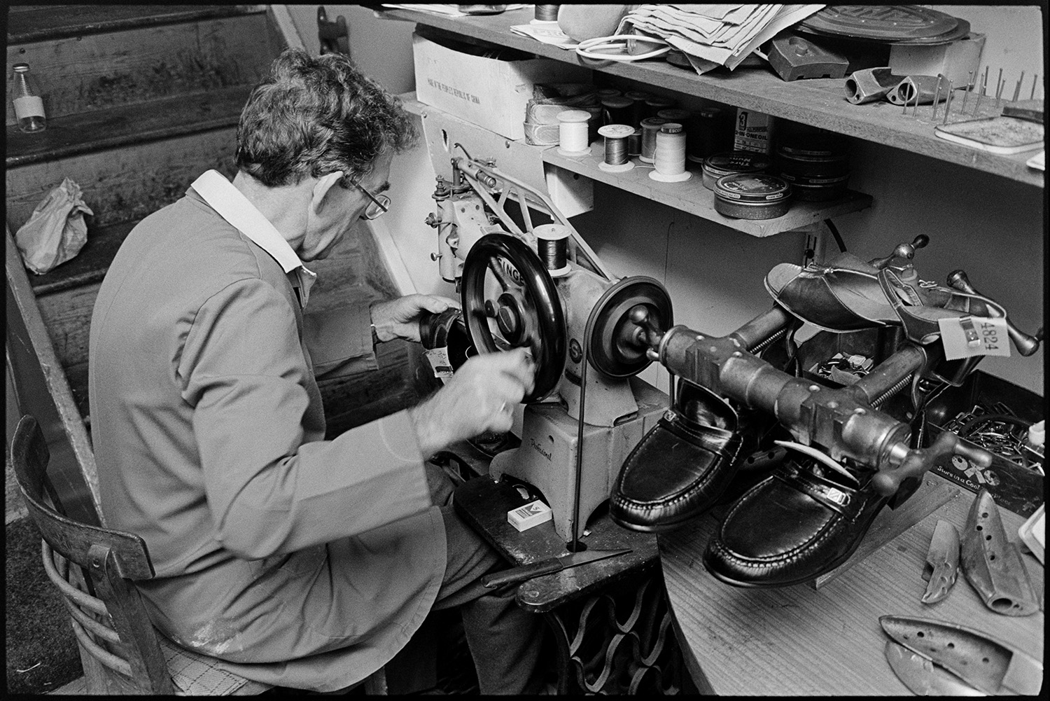 Cobbler mending shoes, interior of workshop, shoe displays. Customers collecting repairs. 
[A man mending shows in his cobbler's workshop in Mill Street, Bideford. He is using a Singer sewing machine.]