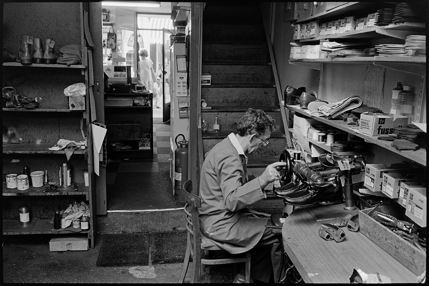 Cobbler mending shoes, interior of workshop, shoe displays. Customers collecting repairs. 
[A man mending shows in his cobbler's workshop in Mill Street, Bideford. He is using a Singer sewing machine. Shelves with various items are visible and the shop front can be seen through an open doorway.]