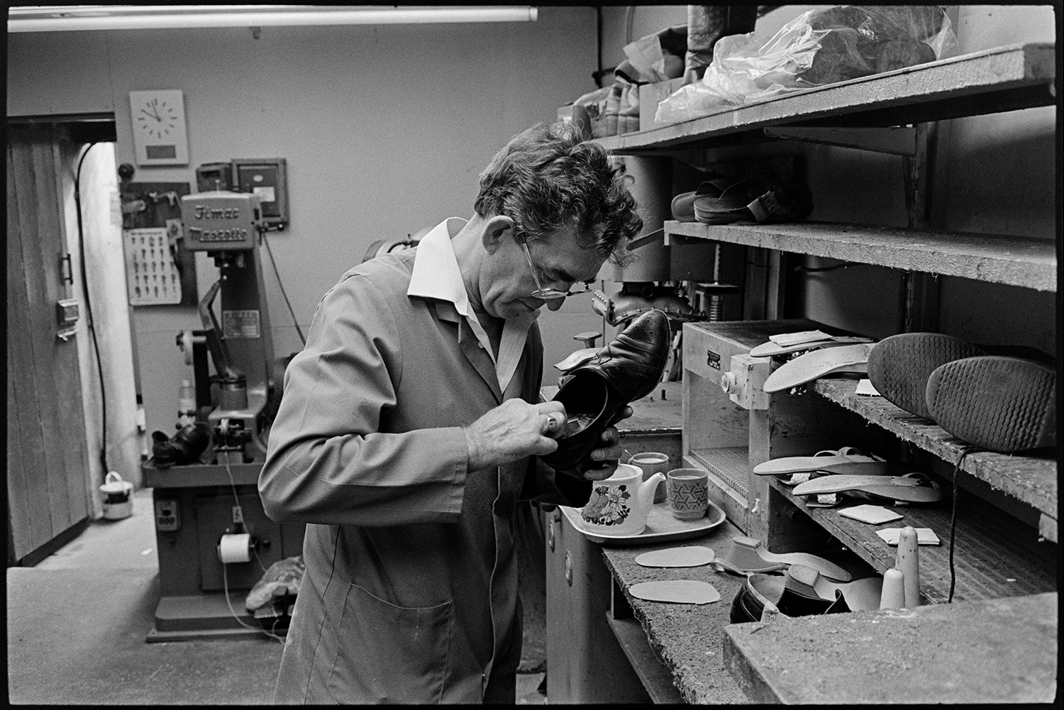 Cobbler mending shoes, interior of workshop, shoe displays. Customers collecting repairs. 
[A man mending a shoe in his cobbler's workshop in Mill Street, Bideford. Shoes can be seen on the shelves in front of him, as well as a tray with a teapot and mugs. A large machine is in the background.]