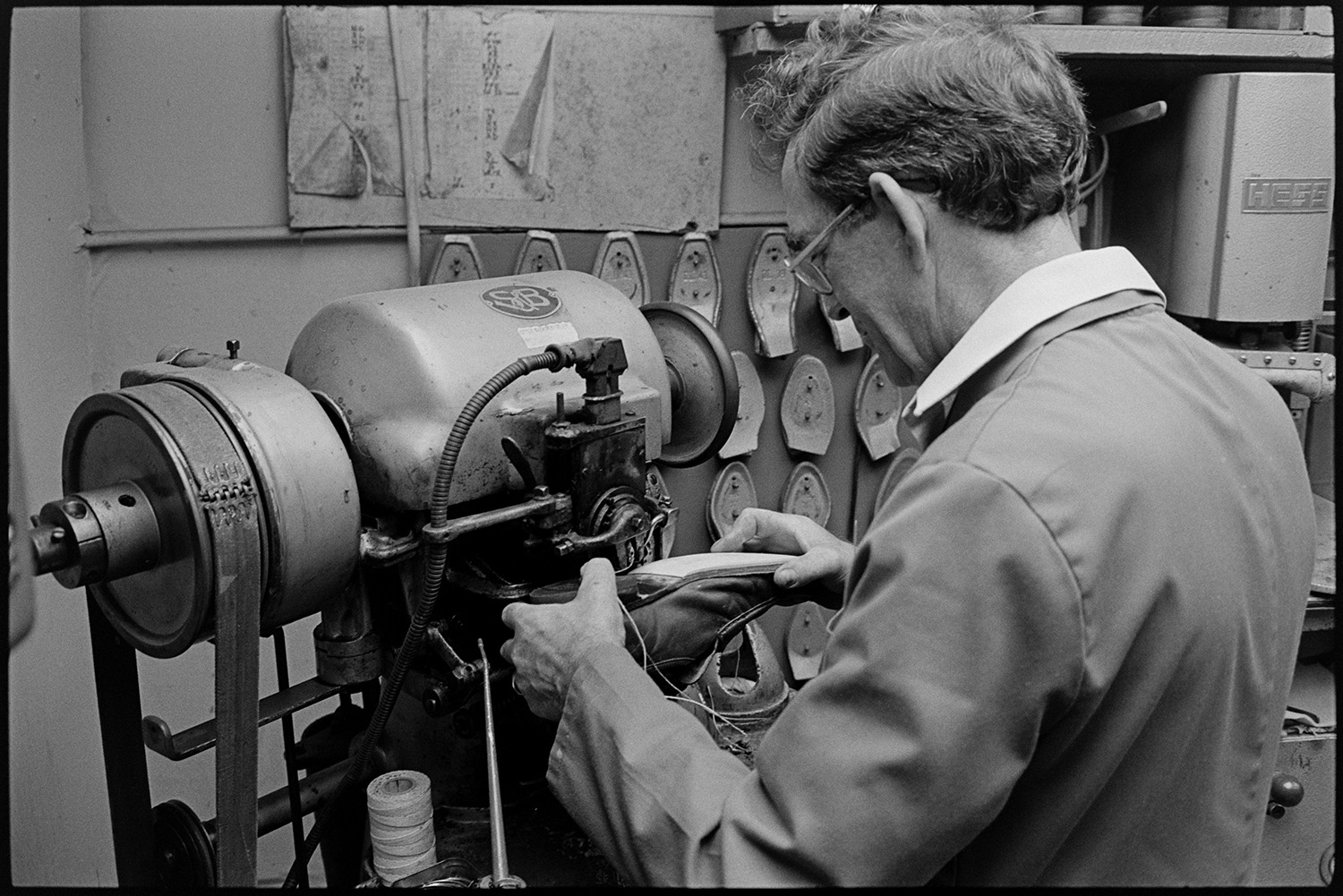 Cobbler mending shoes, interior of workshop, shoe displays. Customers collecting repairs. 
[A man repairing the sole of a shoe using a machine in his cobbler's workshop in Mill Street, Bideford.  Various moulds are hung up on the wall in the background.]