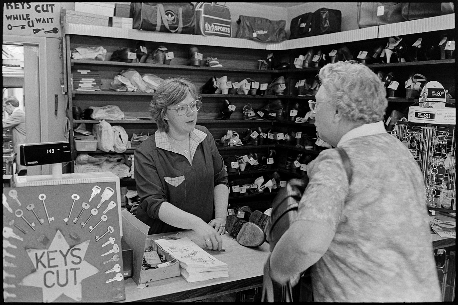 Cobbler mending shoes, interior of workshop, shoe displays. Customers collecting repairs. 
[A woman collecting her repaired shoes from the counter of a cobbler's shop in Mill Street, Bideford. Other shoes waiting to be collected are on the shelves behind the counter and an advert for key cutting is in front of the till.]