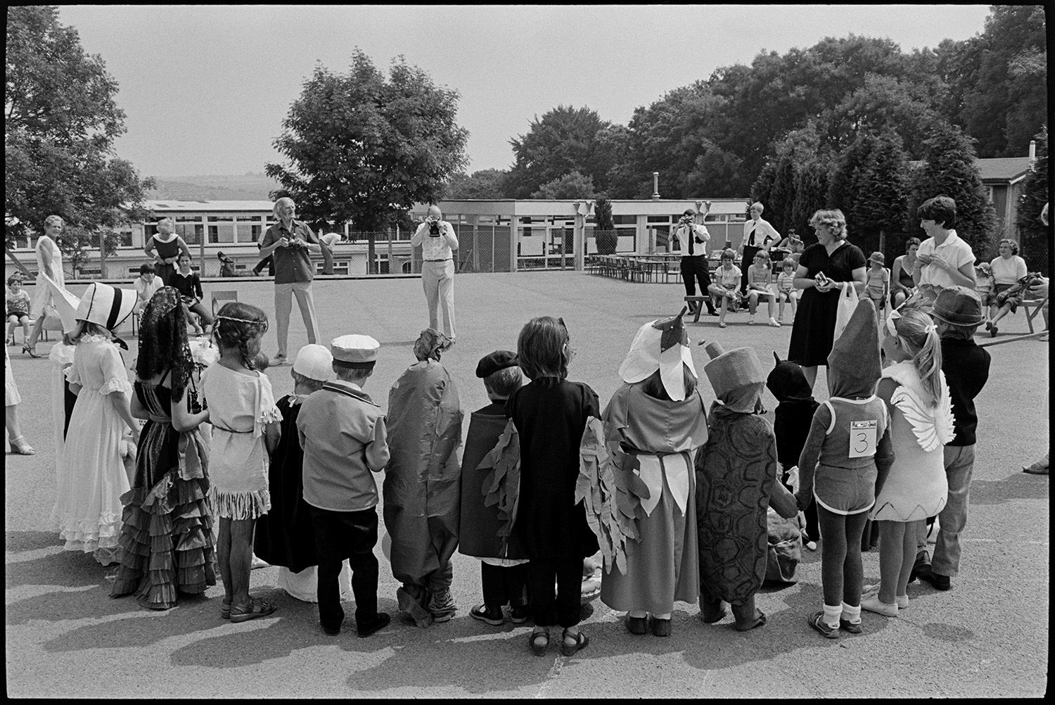 School fete, fancy dress parade mothers and children. 
[People taking photographs of children lined up in their fancy dress costumes at a fancy dress parade at the school fete of Bideford School in the playground.]