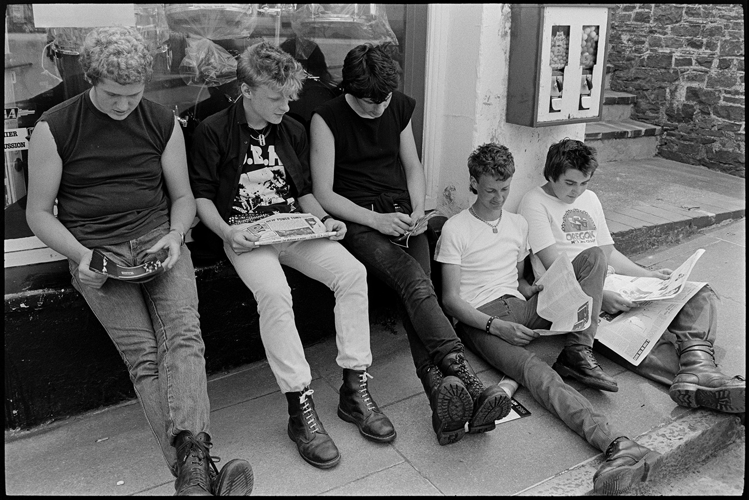 Youths, young men sitting on pavement, clothes, jeans, boots, Doc Martens. 
[Young men sat on a pavement and window sill of a shop front, reading newspapers, at the top of the town in Bideford. Some of them are wearing Doc Marten boots.]