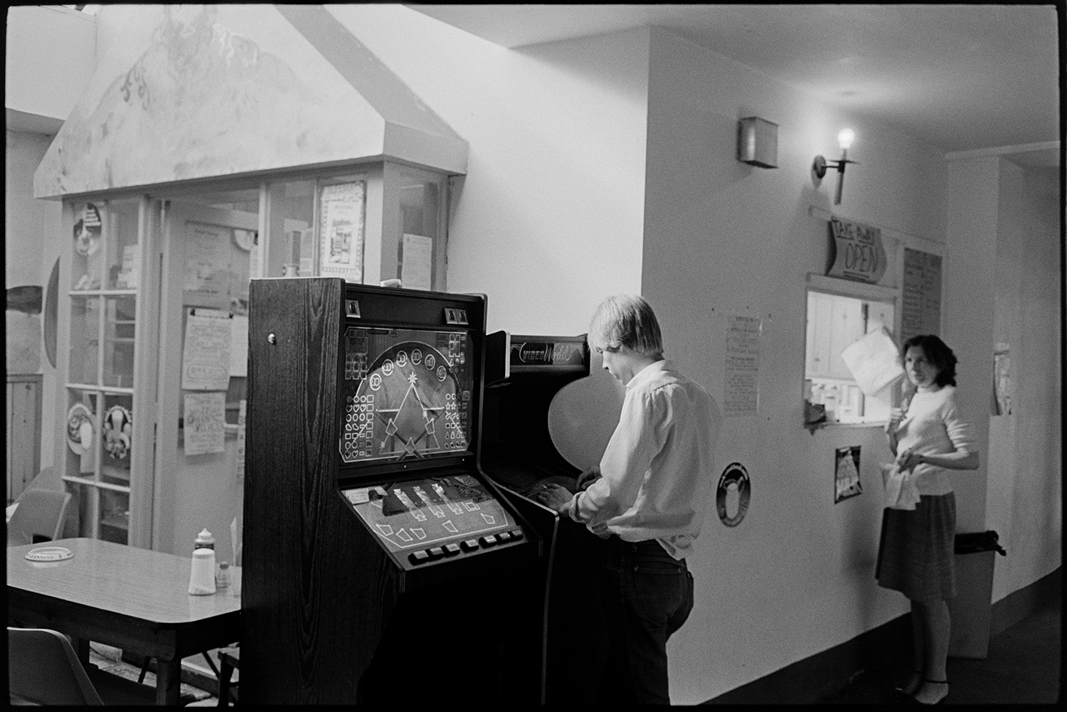 Tea room in covered market. Fruit Machine. 
[A man playing on a fruit machine by a café in Bideford. A woman is waiting for her take away order from the café at a serving hatch in the background.]