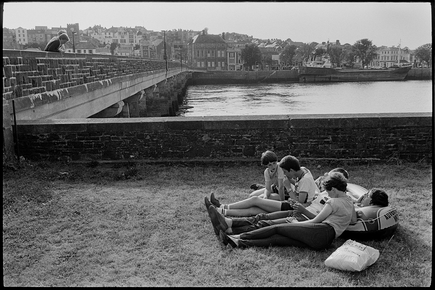 Youths lying beside river on grass, bridge behind. 
[Boys sat on grass beside the River Torridge at Bideford. One of them is resting on an inflatable dinghy or boat. Bideford Long Bridge, also known as Bideford Old Bridge, can be seen in the background along with houses on the opposite side of the river.]