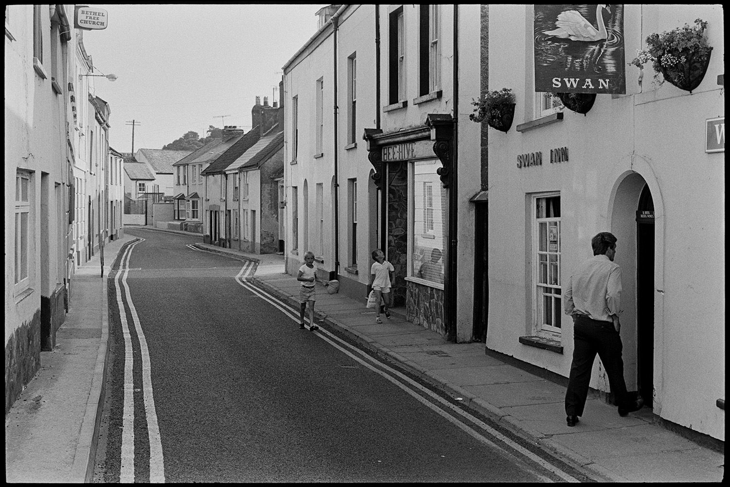 Street scene, public house. 
[A man walking into the Swan Inn at East the Water, Bideford. Two children are walking past the shop front of the 'Beehive' further along the street. Houses and a sign for Bethel Free Church are also visible on the street.]