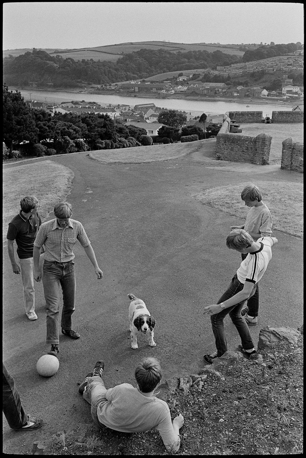 Youths playing football, swings in park, with dog. 
[Young men and boys playing football at Chudleigh Fort overlooking the River Torridge and Bideford. A dog is with them.]