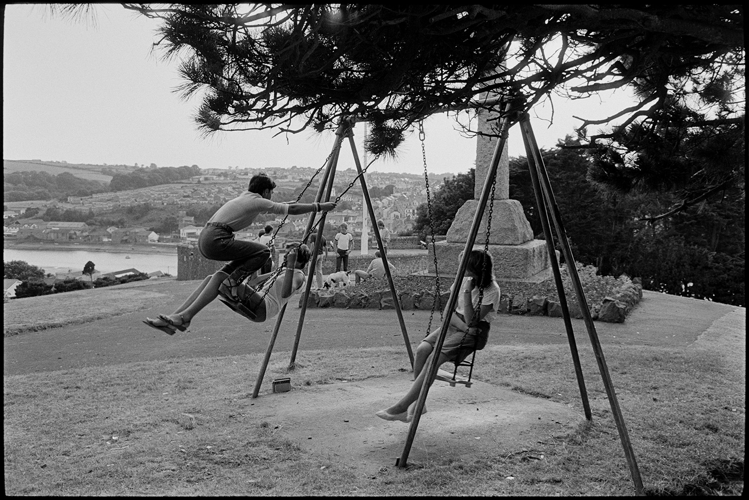 Youths playing football, swings in park, with dog. 
[Young men and women on swings at Chudleigh Fort by the war memorial. The town of Bideford and the River Torridge can be seen in the background.]