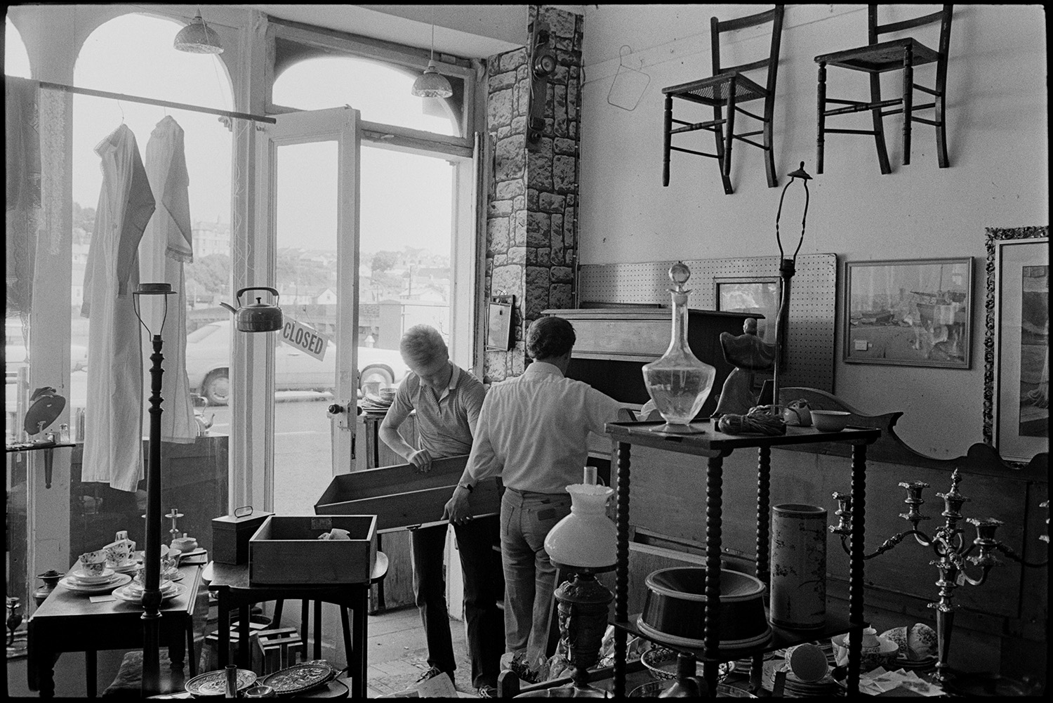 Two men inside Long Johns' Treasure Chest Antique shop on Bideford Quay. They are looking at a chest of drawers. Various items, including tables, china, an oil lamp, a decanter, paintings and chairs hung up on the wall can be seen in the shop.
