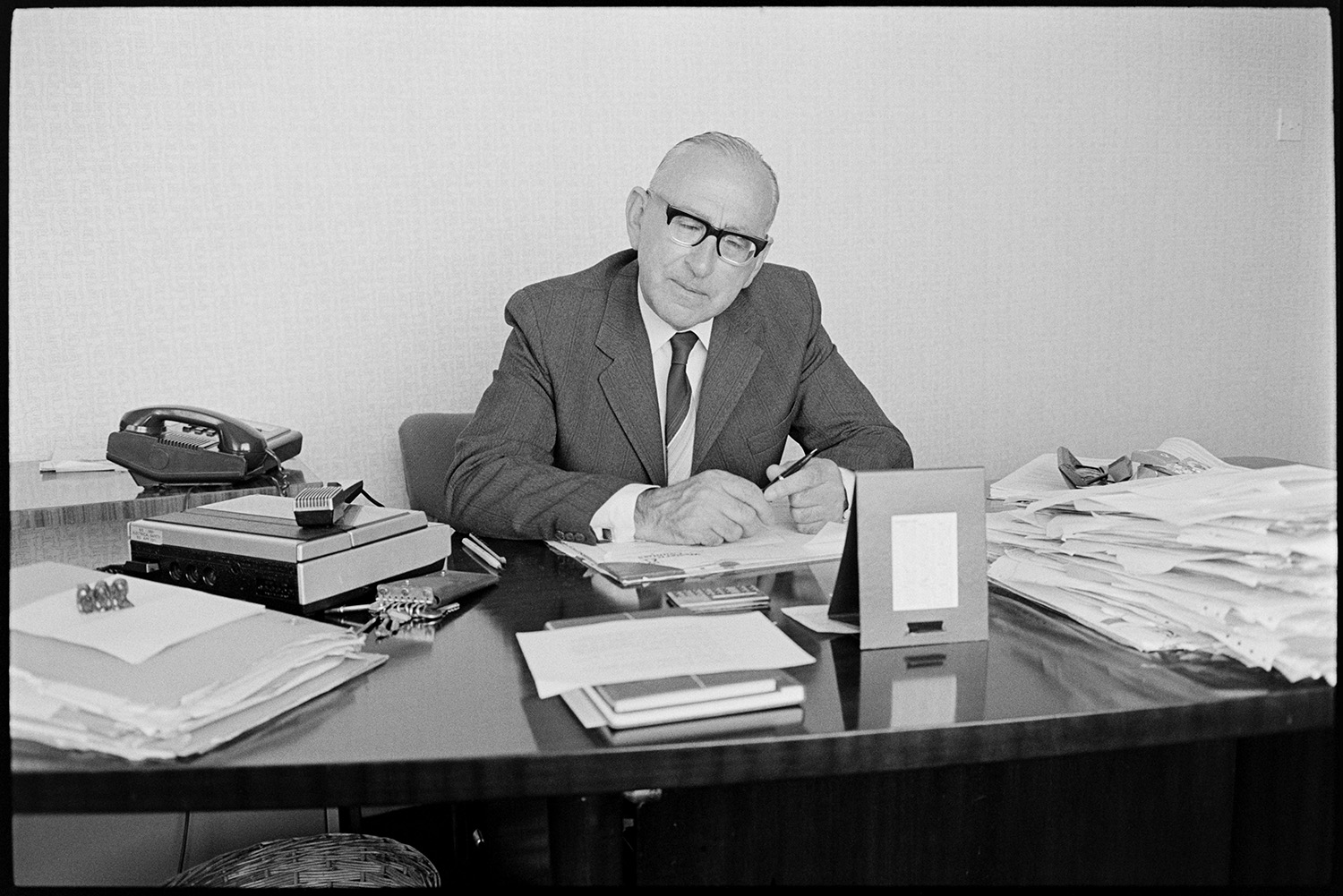 Manager at his desk, Insurance firm?? Mayor of Bideford chatting to photographer. 
[A man sat at a desk in an office, possibly at an Insurance Firm, in Bideford. Various papers, a telephone and a calculator can be seen on the desk.]