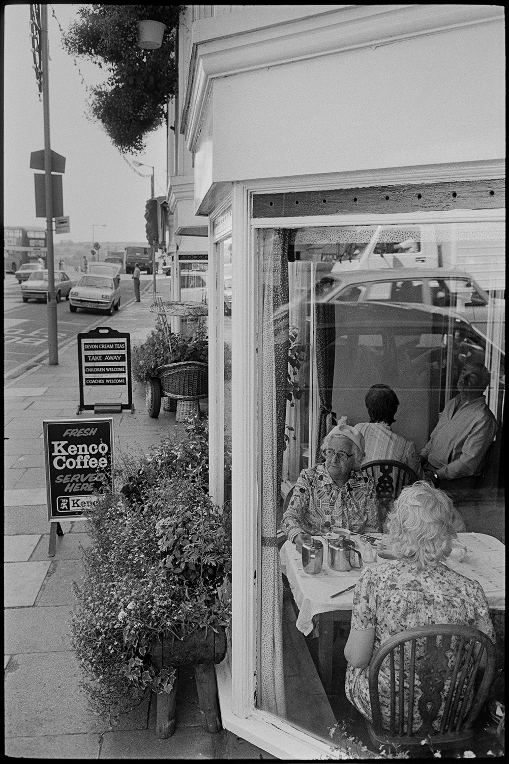 Street scene, women in café, holidaymakers at table outside. 
[People sat at tables in The Rose of Torridge tea room at Bideford Quay. Two women are sat by the window. Signs advertising coffee and window boxes can be seen on the pavement outside.]