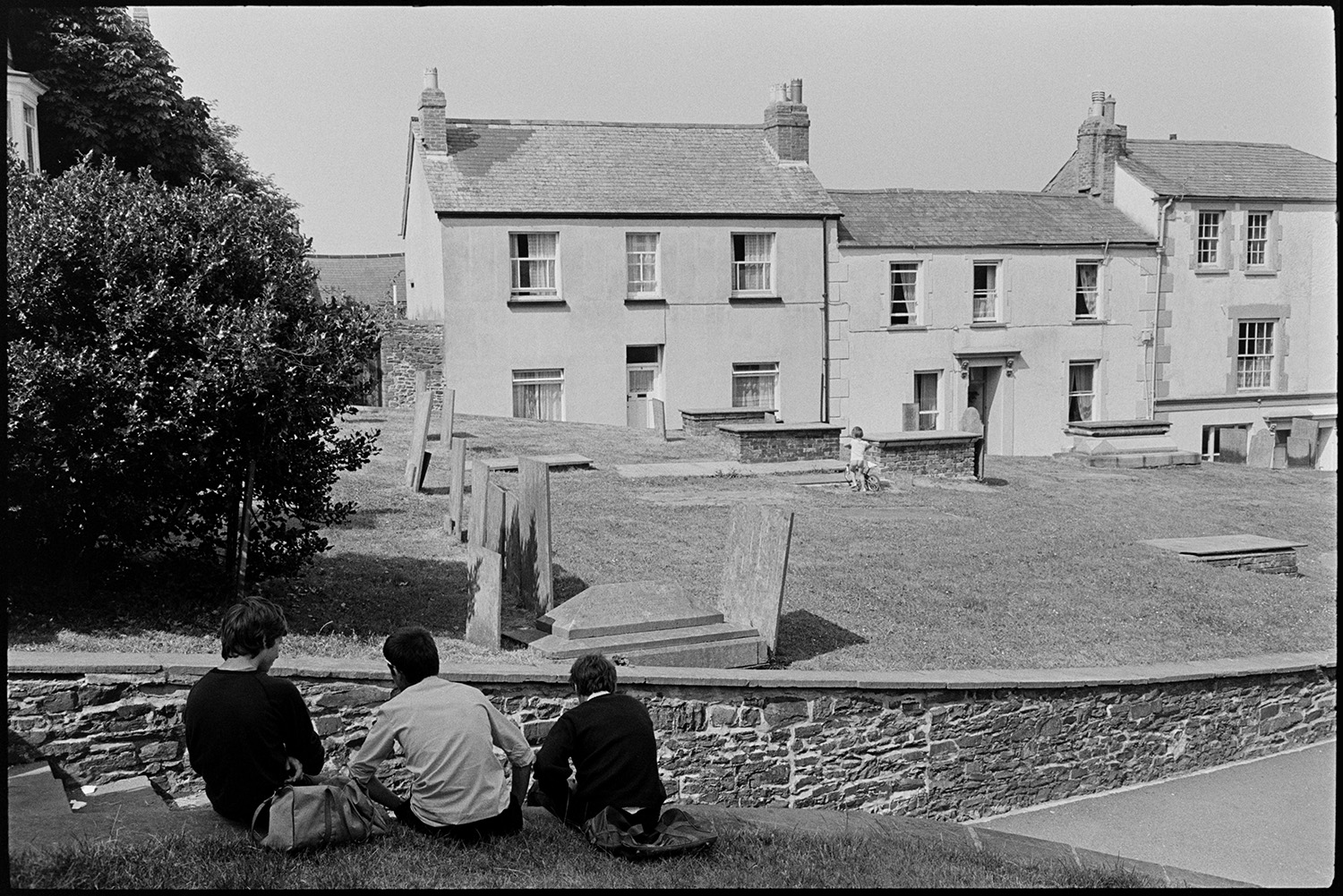 Street scenes, graveyard with boys sitting on wall. 
[Three boys with bags sitting on a wall running alongside a path through Bideford Churchyard. A young child can be seen playing with a bicycle amongst the gravestones and terraced houses are visible opposite the graveyard.]