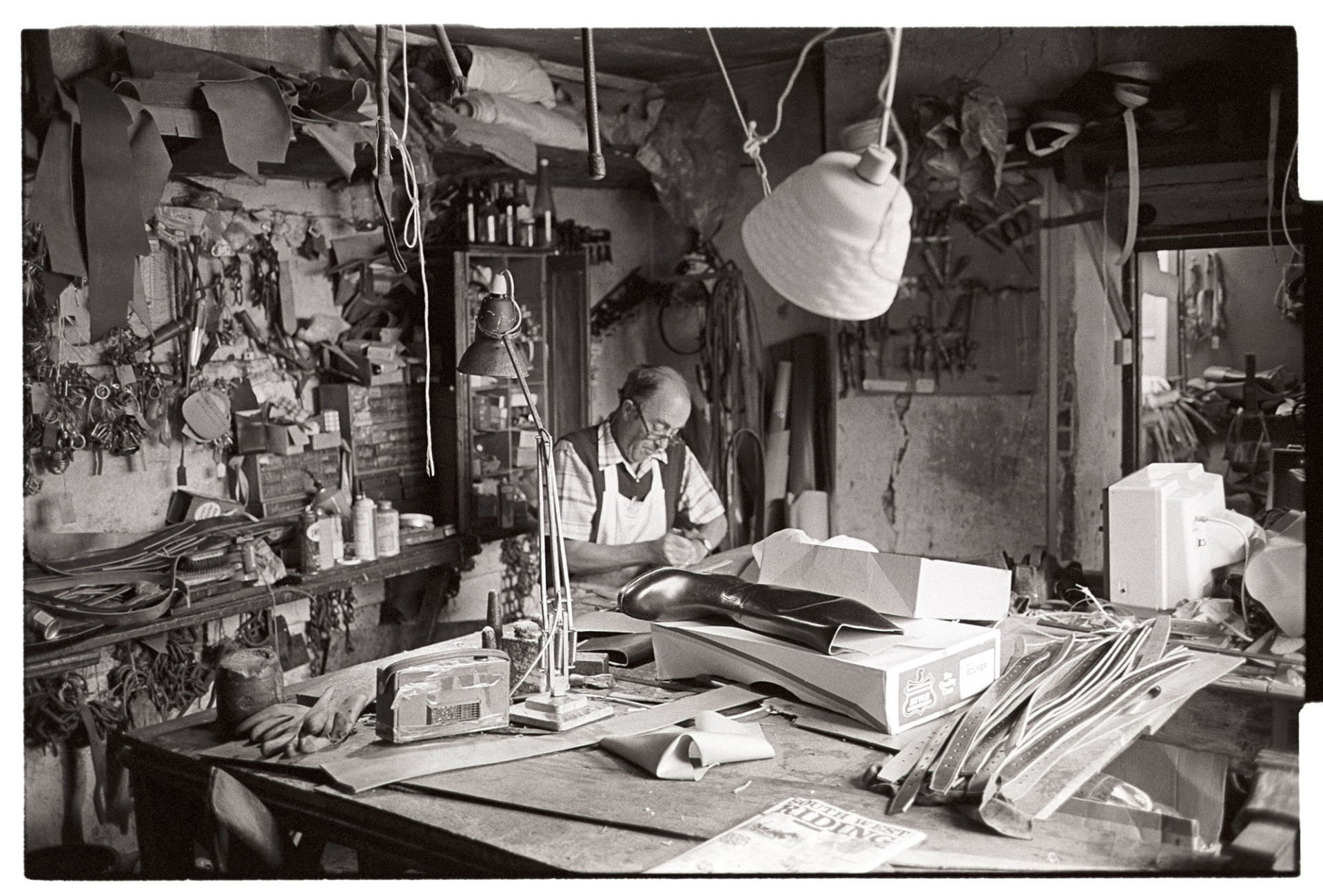 Saddler in his workshop, tools, materials, goods on display. <br />
[Derek Johns, a saddler, sat at a bench in his workshop at Buttgarten Street, Bideford. A lamp and radio are also on the workbench. Various goods and tools are on display, including a boot, belts or straps and gloves.]
