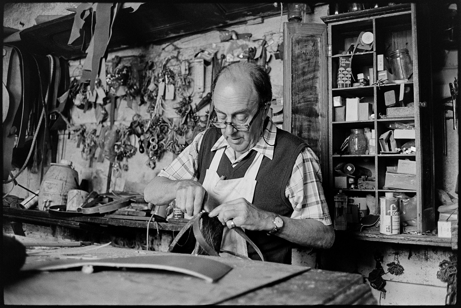 Saddler in his workshop, tools materials, goods on display. 
[Derek Johns, a saddler, making leather goods in his workshop in Buttgarden Street, Bideford. Various tools are hung up on the wall behind him.]