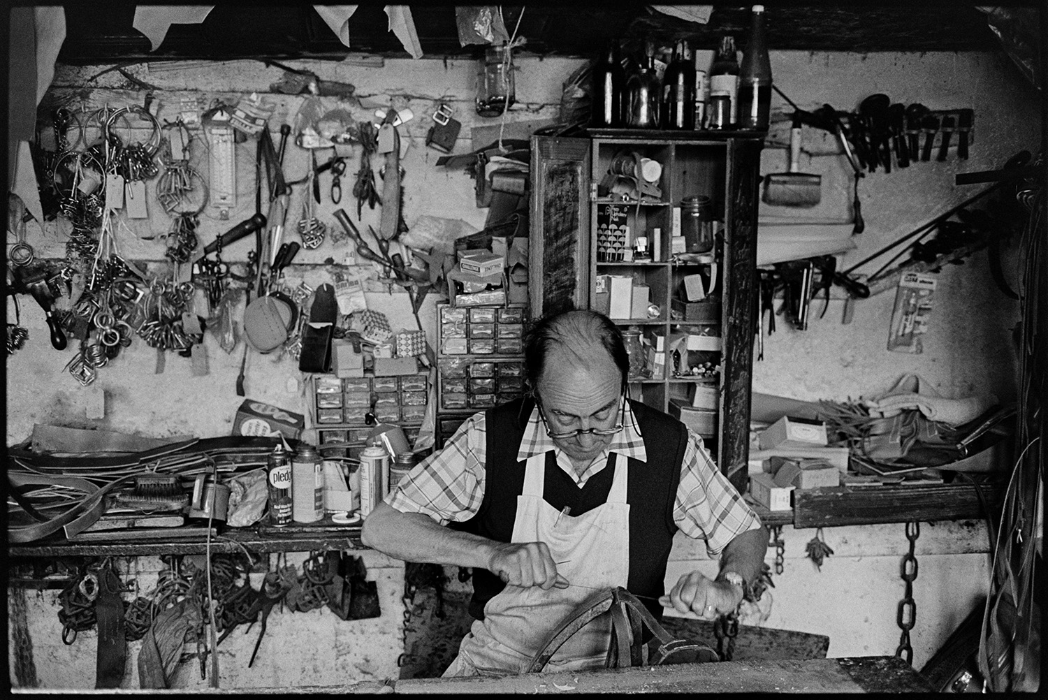 Saddler in his workshop, tools materials, goods on display. 
[Derek Johns, a saddler, making leather goods in his workshop in Buttgarden Street, Bideford. Various tools are hung up on the wall behind him by shelves and small filling drawers.]