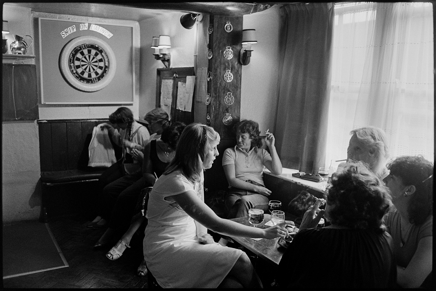 People drinking and playing darts in pub. 
[A group of women drinking and smoking in the Blacksmiths Arms pub at East the Water, Bideford. A dartboard can be seen on the all in the background.]