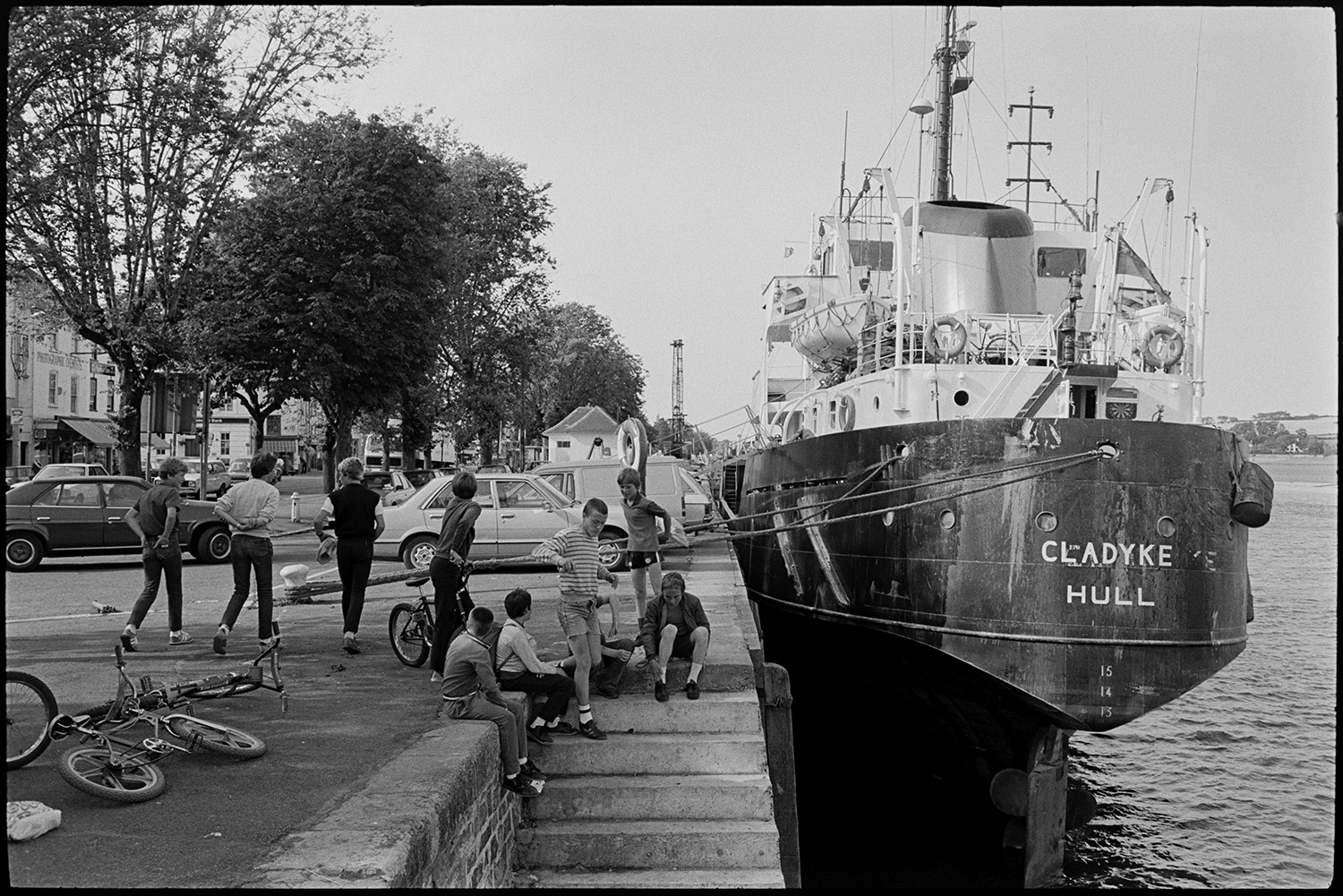 Ship at quayside, children with bicycles. 
[Children playing by steps leading down from Bideford Quay to the River Torridge. The ship, Cladyke, is moored beside the quay. Parked cars and children's bicycles can be seen on the quay.]