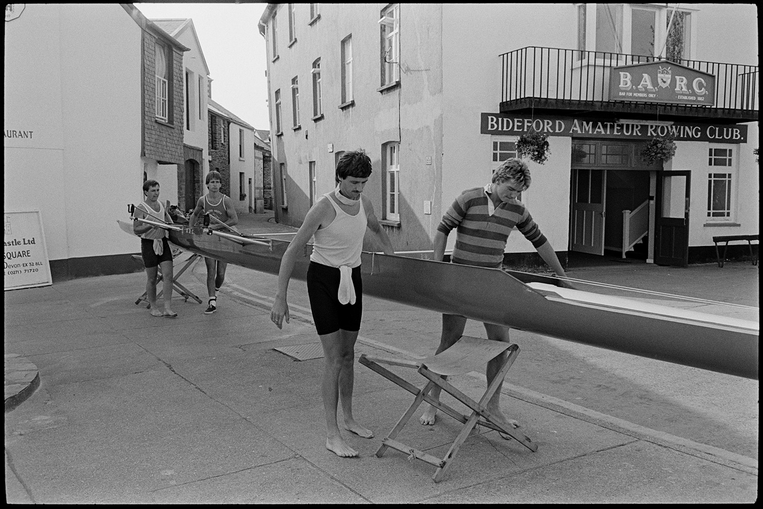 Oarsmen launching racing boat. 
[Four men carrying a racing boat to launch on the River Torridge at Bideford Quay. They are outside Bideford Amateur Rowing Club.]